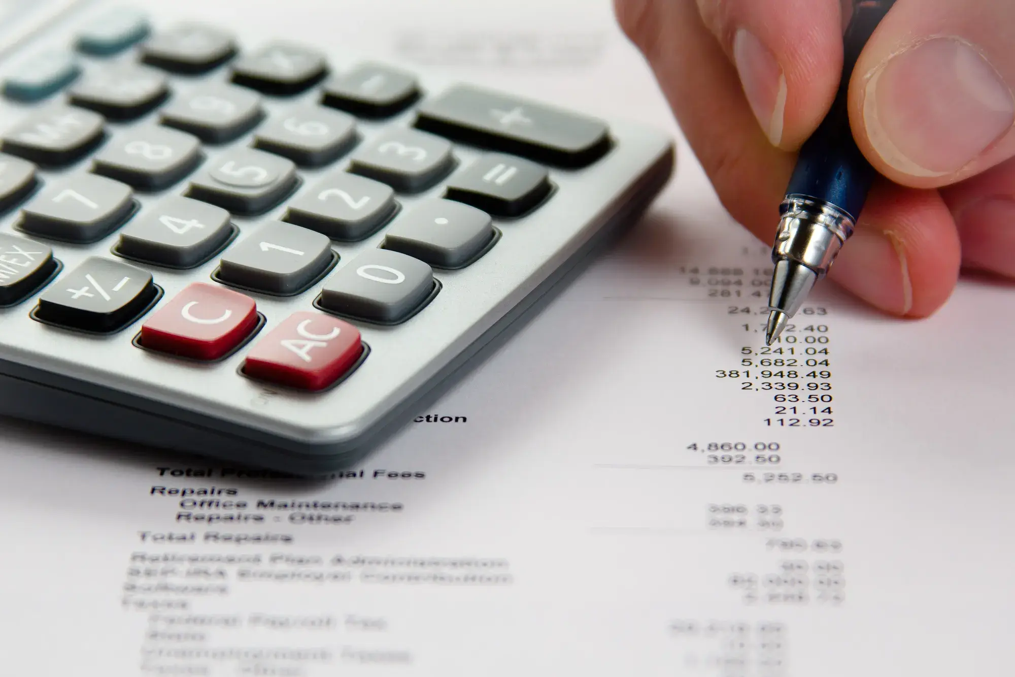 A hand with a pen working through printed financial figures beside a calculator