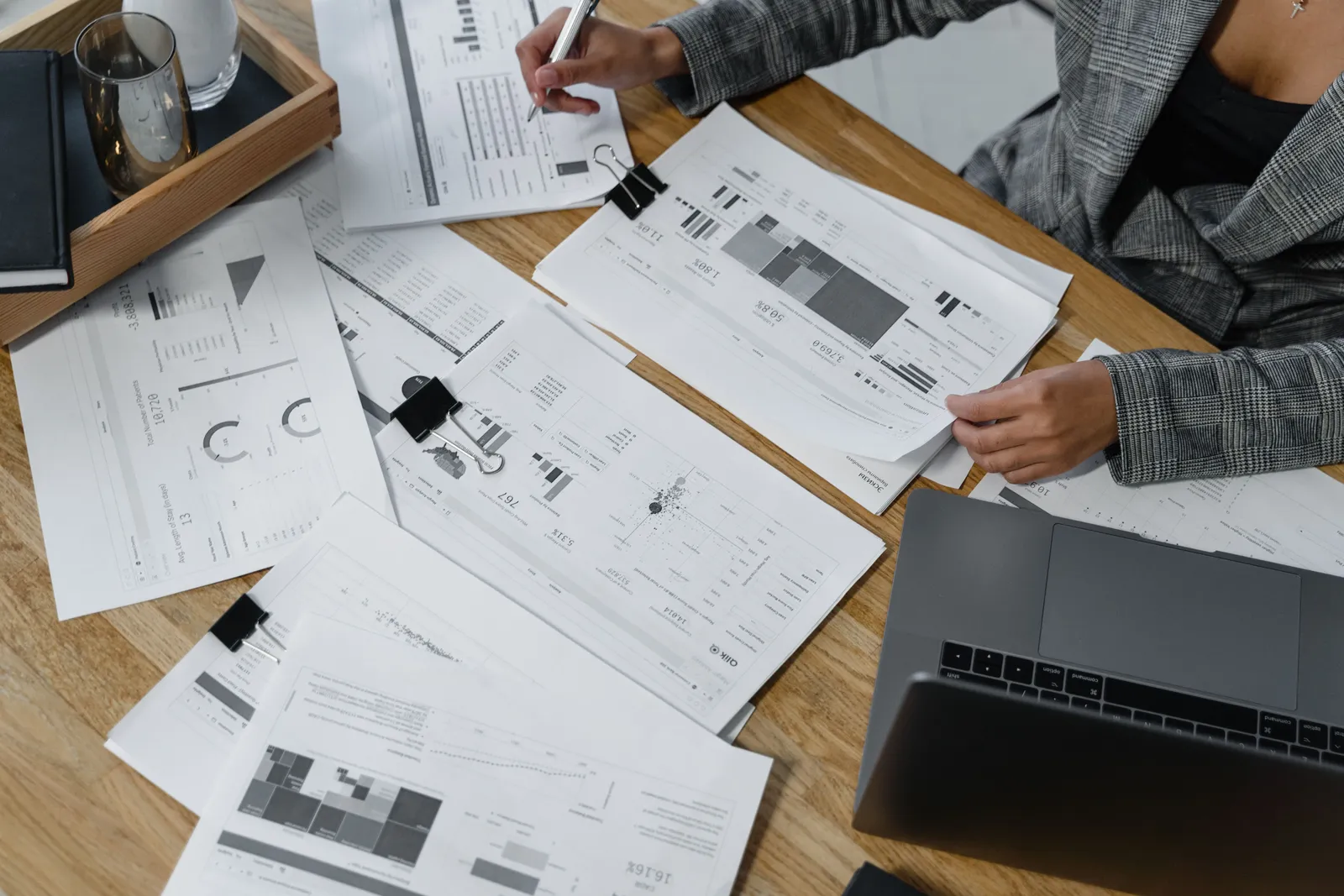 A person reviewing printed financial charts and reports spread across a desk beside a laptop